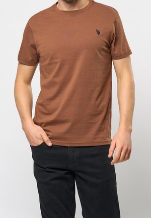 Uomo che indossa una maglietta marrone a maniche corte con un piccolo logo ricamato blu navy sul petto e pantaloni neri, una mano in tasca.