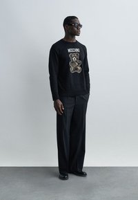 Άνδρας που φοράει μαύρο πουλόβερ Moschino με σχέδιο αρκουδάκι, μαύρο φαρδύ παντελόνι, μαύρα παπούτσια και σκούρα γυαλιά ηλίου, στέκεται μπροστά σε απλό φόντο.