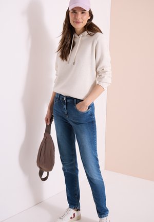 Femme portant un sweat à capuche beige clair, un jean bleu, des baskets blanches et une casquette rose, tenant un sac marron dans sa main gauche.