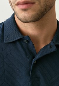 Cămașă polo bleu închis cu model texturat în herringbone, având guler și plachetă cu două butoane; material moale și o strălucire subtilă.