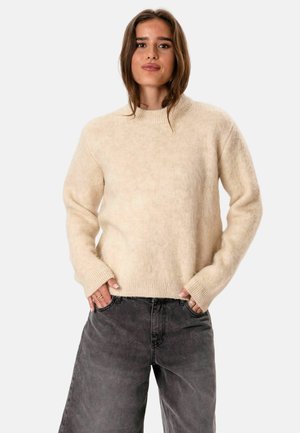 Femme aux cheveux bruns porte un pull duveteux couleur crème et un jean ample gris foncé, debout les mains dans les poches devant un fond uni.