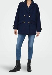 Donna che indossa un cappotto a doppiopetto blu navy con bottoni dorati, jeans skinny azzurri e stivali neri alla caviglia, in piedi su uno sfondo bianco.