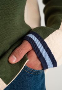 Žalias megztinis su kreminiais ir mėlynais akcentais, turintis juostuotas rankoves. Rankos ilsisi mėlynųjų džinsų kišenėje. Matomos tekstūruotos audinio detalės.