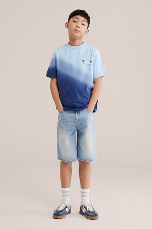 Chlapec stojaci s rukami v vreckách, oblečený v modrom ombré tričku, svetlých rifľových šortkách, bielych ponožkách a modrých teniskách na jednoduchom pozadí.