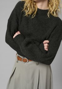 Blonde Person mit verschränkten Armen, die einen schwarzen Rippensweater, beige Hosen und einen braunen Gürtel mit einer silbernen Schnalle trägt, vor einem grauen Hintergrund.