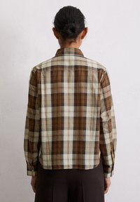 Chemise à carreaux marron et crème avec des manches longues, des poignets boutonnés, un col classique et une coupe décontractée. La texture du tissu semble légère.