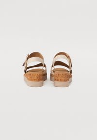 Beige strappy sandals with a textured brown wedge heel. Features an intricate floral accent on the side and a smooth inner lining.