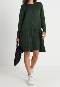 Vestido verde oscuro con mangas largas y una falda fruncida. Presenta lunares blancos, un escote redondo y una tela texturizada. Se lleva con zapatillas blancas y un bolso negro.