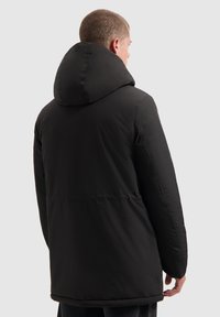 Zwarte gewatteerde jas met capuchon, gladde stof, iets baggy pasvorm, naadtoeslagen en een langere achterkant voor extra bedekking.