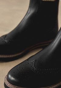 Mustast nahast Chelsea-saapad brogue-detailidega, millel on sile viimistlus, ümar varvas ja kontrastne hele tallaosa.