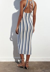 Robe en tricot rayée avec des rayures verticales bleues et crème, dos ouvert avec un détail noué, longueur mi-mollet, associée à des talons beiges à lanières.
