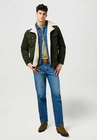 Grüne Cordjacke mit weißem Pelzkragen, darunter ein Jeanshemd, ein olivfarbenes T-Shirt, blaue Jeans und braune westernstile Stiefel.