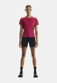 Červené sportovní tričko s krátkým rukávem a elegantním designem, doplněné černými šortkami. Materiál vypadá lehce a prodyšně, ideální pro sport.