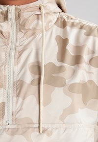 Chaqueta con capucha ligera en patrón de camuflaje beige y marrón, con cremallera frontal y cordón en el cuello para un ajuste regulable.