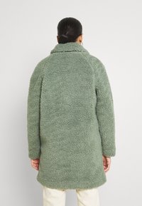 Faux fur, överdimensionerad grön kappa med hög krage och långa ärmar, texturerad tyg och rak avslutning. Bakvy visas.