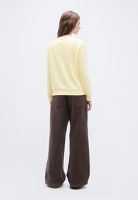 Γυναίκα με την πλάτη, φορώντας ανοιχτόκίτρινο πουλόβερ και σκούρο καφέ παντελόνι με φαρδιά πόδια, στέκεται μπροστά σε απλό ανοιχτό φόντο.