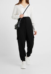 Pantaloni cargo neri con tasche laterali e vita con cordino, abbinati a una maglietta bianca a maniche lunghe e scarpe da ginnastica bianche, con una piccola borsa nera.