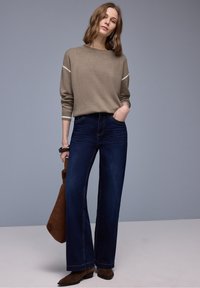 Femme portant un pull taupe et un jean large bleu foncé, tenant un sac en daim marron, debout devant un fond gris uni.