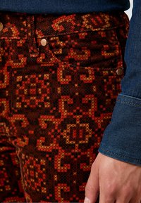 Hand som vilar bredvid en närbild av röda och orange mönstrade corduroybyxor och en ärm av en blå denimskjorta.