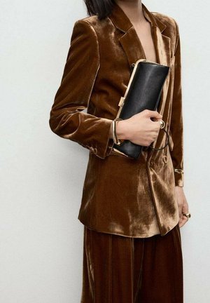 Personne portant un costume en velours marron tenant une pochette noire, portant des bracelets et des bagues en or, recadrée au niveau du cou et des genoux.