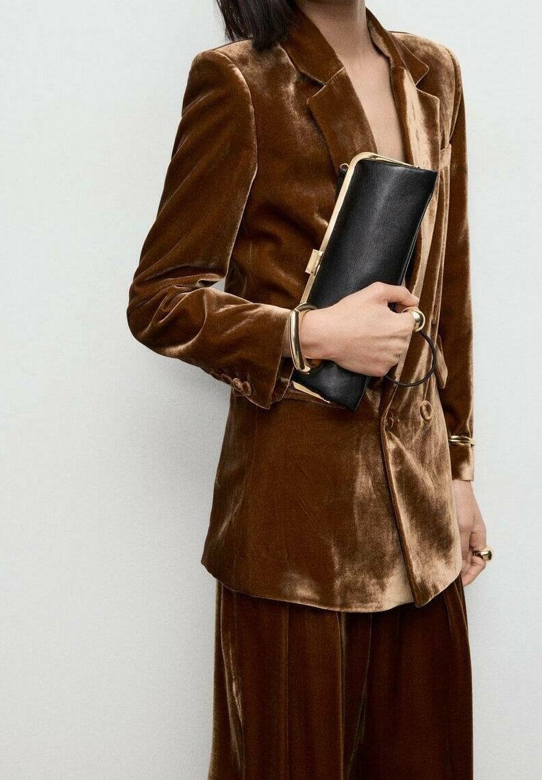 Personne portant un costume en velours marron tenant une pochette noire, portant des bracelets et des bagues en or, recadrée au niveau du cou et des genoux.