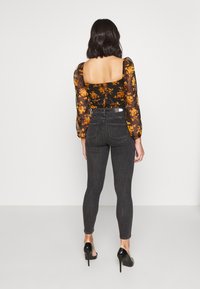 Blusa negra transparente de manga larga con estampado floral naranja, combinada con jeans ajustados de cintura alta en gris oscuro y zapatos de tacón negro.