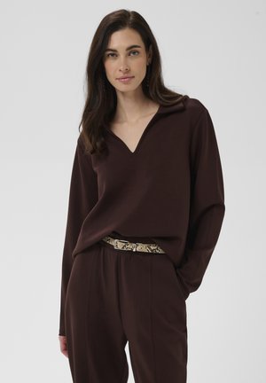 Femme aux longs cheveux foncés portant un chemisier marron foncé en V et un pantalon assorti avec une ceinture à motif peau de serpent.