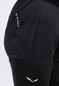 Κοντινό πλάνο μαύρου παντελονιού από ελαστικό ύφασμα με φερμουάρ στην τσέπη και λευκό κείμενο "DURASTRETCH" και λογότυπο στο μηρό.