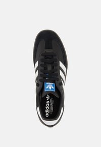 Černá teniska s semišovým a syntetickým svrškem, bílými pruhy, černými tkaničkami a polstrovaným límcem. Na jazyku je modré logo Adidas.