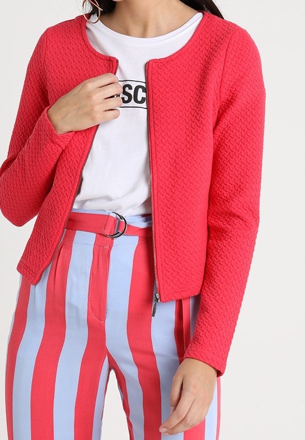 Femme portant une veste zippée rouge vif texturée par-dessus un t-shirt blanc à motif, associée à un pantalon taille haute à rayures verticales rouges et bleu clair.
