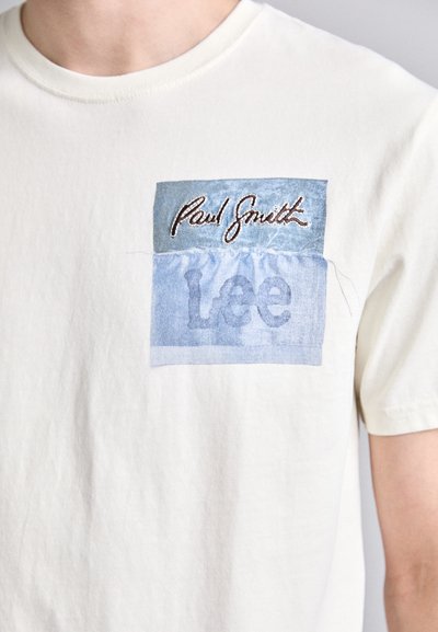 Balta medvilninė marškinėlių su mėlynu stačiakampiu pamušalu, kuriame siuvinėti žodžiai "Paul Smith" virš "Lee." Minkšta tekstūra, laisvalaikio dizainas.