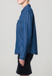 Camicia in denim di colore blu scuro, con colletto, chiusura frontale con bottoni e una tasca sul petto. Maniche lunghe con polsini, vestibilità rilassata.