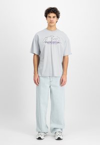 Svijetlosivi oversized pamučni majica s natpisom "ALPHA STUDIO EDITION". Uskladena s plavim, širokim trapericama i bijelim tenisicama.