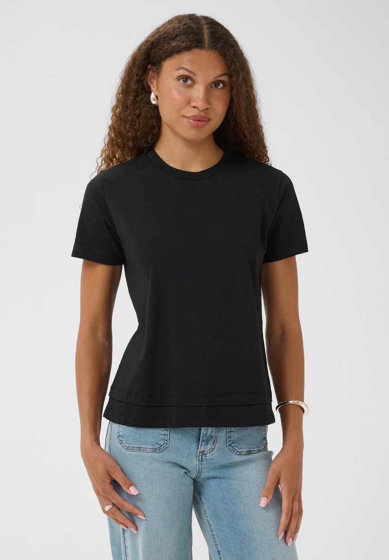 Femme aux cheveux bouclés portant un t-shirt noir uni à manches courtes et un jean bleu clair, debout devant un fond uni.