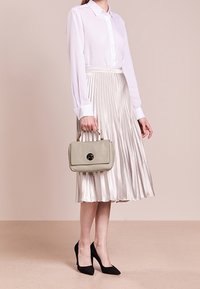 Nainen, jolla on päällään valkoinen paita ja hopeanvärinen pleated-mittapituinen hame, pitää beigeä käsilaukkua ja mustia korkokenkiä, seisoo beigeä taustaa vasten.