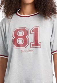 Harmaa lyhythihainen T-paita, jossa on punainen ja valkoinen ribattu kaulus. Sisältää rohkean punaisen "81" -grafiikan ja "Guess Jeans" -tekstin painettuna alapuolelle.