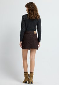 Vrouw met krullend haar, gekleed in een donkere trui, een bruine denim minirok en beige suède enkelhoge laarzen met hakken, staand met haar gezicht naar een effen witte achtergrond.