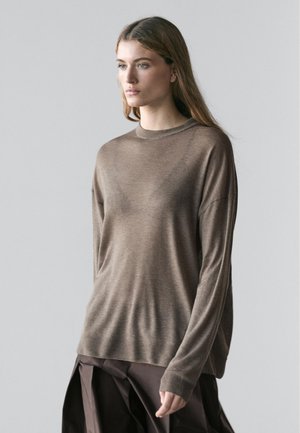 Woman with long hair wearing a loose brown sweater and dark brown skirt, standing against a plain light gray background.