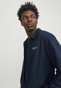 Tamno plavi zip-up jakna s kragnom, s logotipom na lijevoj strani prsnog koša. Glatka tekstura, lagani dizajn i bočni džepovi.