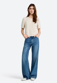 Blouse courte à manches courtes en crème avec des manches bouffantes, associée à un jean en denim bleu à jambe large. Le mannequin se tient avec les mains dans les poches, portant des chaussures minimalistes.