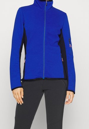 Blauer, strukturierter Reißverschluss-Pullover mit schwarzen Seitenpaneelen und Reißverschlusstaschen, getragen zu einer Person, die dunkle, eng anliegende Hosen trägt und steht.