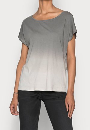 Vrouw die een korte mouw ombre top draagt die vervaagt van grijs naar wit en zwarte broek, staand tegen een effen achtergrond.