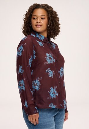 Kvinde med krøllet hår iført en vinrød langærmet skjorte med blå blomsterprint og blå jeans, kigger til siden.
