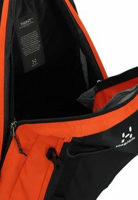 Mochila con un material exterior negro y un interior naranja brillante. Cuenta con un cierre de cremallera y un bolsillo interior. Etiqueta de la marca visible.