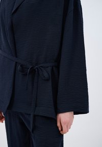 Navy blue textured robe featuring wide sleeves, a tie belt, and a relaxed fit. The fabric has a crinkled appearance.