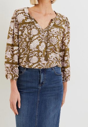 Vrouw draagt een olijfgroene bloes met bloemenprint en kwastjes en een blauwe denim rok met hoge taille, staand tegen een effen lichte achtergrond.