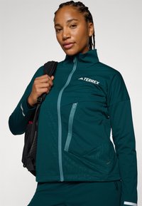 Veste zippée turquoise avec un col haut, présentant un motif texturé sur le devant, un design élégant et un logo TERREX sur la poitrine.