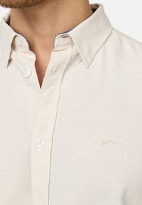 Camisa de botones beige claro con un collar estructurado, que presenta un sutil logo bordado y un ribete azul contrastante en el interior del collar.