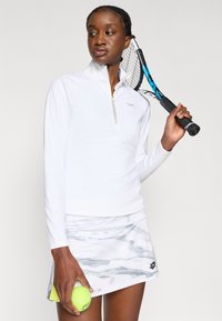 Baltas sportinis marškinėliai su pusiau užtrauktuku ir ilgomis rankovėmis, derinami su balta ir pilka kamufliažo rašto teniso sijonu. Laikydama teniso kamuoliuką ir raketę.