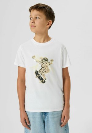 Garçon en t-shirt blanc avec un graphique de skateur, portant un jean bleu clair, regardant sur le côté sur un fond clair uni.